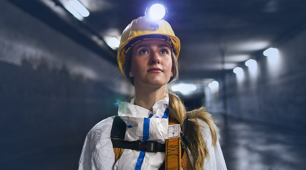 Eine junge Frau mit weißem Overall, Helm und leuchtender Helmlampe steht in einem unterirdischen Kanal und blickt hoffnungsvoll nach vorn.