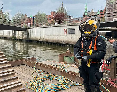 Ein Taucher mit Anzug und Helm steht auf einem Ponton auf dem Wasser in der Hamburger Speicherstadt