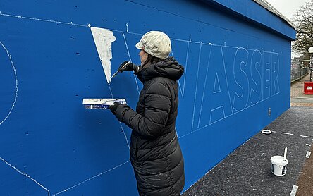Eine Frau schreibt mit weißer Farbe an die blaue Wand des Pumpwerks Hafenstraße.
