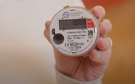 Eine Frau hält einen digitalen Wasserzähler von HAMBURG WASSER hoch. Er ist rund und hat ein Display, auf dem eine Angabe zum Verbrauch erscheint.  