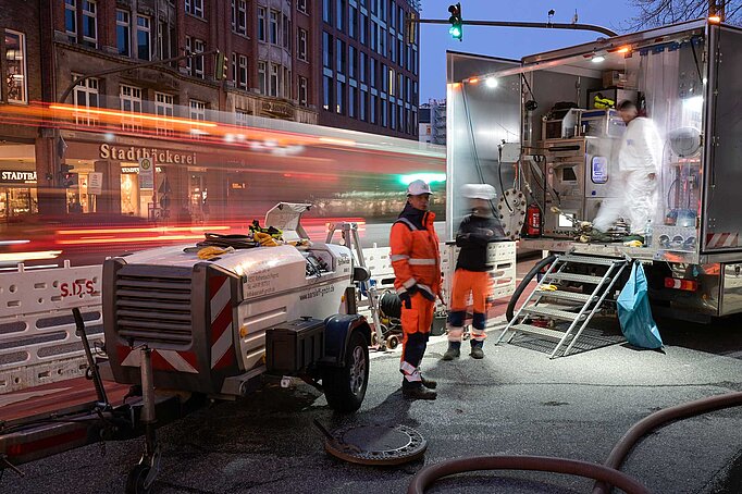 Zwei Arbeiter stehen vor einem Baustellenfahrzeug mit Technik. Es wird langsam dunkel und im Hintergrund sieht man einen Bus fahren. 