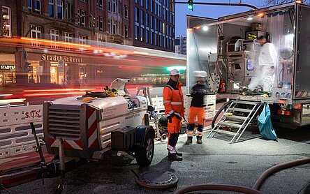 Zwei Arbeiter stehen vor einem Baustellenfahrzeug mit Technik. Es wird langsam dunkel und im Hintergrund sieht man einen Bus fahren. 