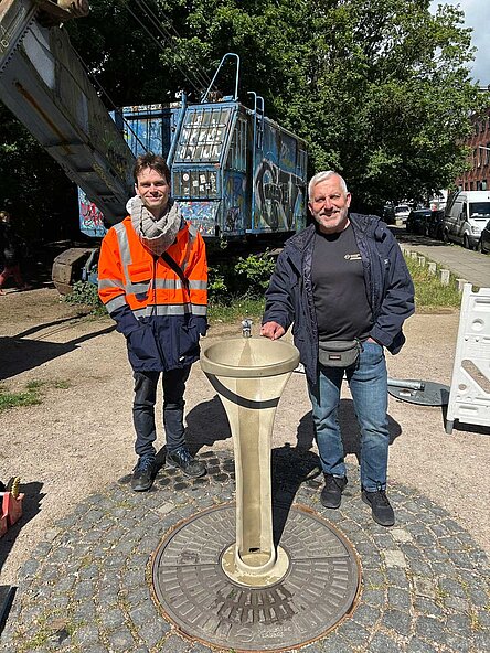Zwei Männer in Arbeitskleidung stehen an einem Brunnen, den sie gerade aufgebaut haben und lächeln in die Kamera. Der Brunnen ist bronze und hat ein schlankes Design, das frische Trinkwasser geht in einem Strahl durch einen Bogen und fließt dann in eine Auffanschale, die direkt über dem Boden ist. Im Hintergrund sieht man einen alten Bagger, der als Industriedenkmal auf dem Platz in Ottensen steht.