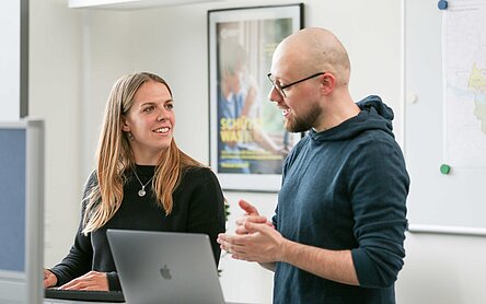 Eine Mitarbeiterin und ein Mitarbeiter disktieren angeregt vor einem Laptop stehend