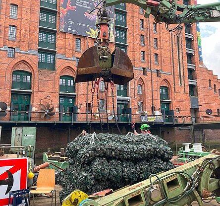 Die Steine in Netzen sehen aus wie Matten. Dahinter steht ein Bagger auf einem Ponton, der die Matten in den Kanal in der Speicherstadt heben soll.