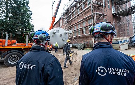 Zwei Ingenieure von HAMBURG WASSER stehen im Vordergrund, im Hintergrund wird gerade ein neuer Filterkessel in das Wasserwerk eingehoben.