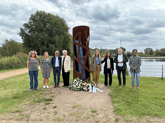 Eine Gruppe von Menschen steht vor dem Mahnmal, das hoch in die Luft ragt und an dessen Fuß Kränze niedergelegt sind.