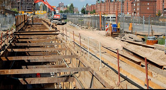 Spundwände stützen eine große Grube, die tief in die Erde ragt. Die Baustelle ist abgesperrt, im Hintergrund sind Häuser zu sehen.