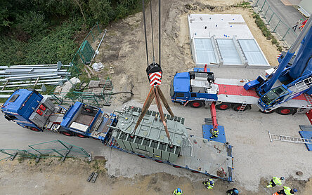Ein blauer Schwerlasttransporter hat einen sehr großen Trafo geladen, der per Kran in das Baufeld für die Wärmepumpe soll. Das Foto ist aus der Vogelperspektive mit einer Drohne entstanden.