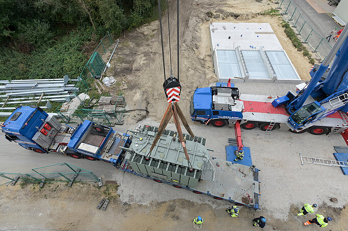 Ein blauer Schwerlasttransporter hat einen sehr großen Trafo geladen, der per Kran in das Baufeld für die Wärmepumpe soll. Das Foto ist aus der Vogelperspektive mit einer Drohne entstanden.