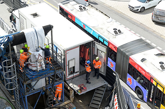 Zu sehen ist ein großer Schlauch, der über ein Gerüst ins Siel eingelassen wird, links, direkt daneben fährt ein Bus vorbei, rechts. Es ist wenig Platz. 