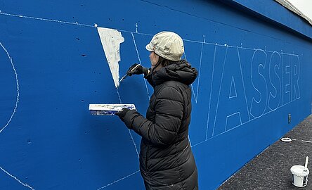 Eine Frau schreibt mit weißer Farbe an die blaue Wand des Pumpwerks Hafenstraße.