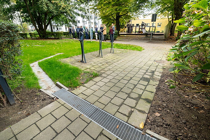 Auf dem Boden sieht man einen geflasterten Weg, in dem ein Wasserweg läuft, der durch ein Gitter geschützt wird. Der Wasserweg läuft in eine Mulde aus Gras