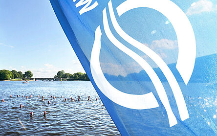 Eine Fahne mit dem Logo von HAMBURG WASSER vor der Alster, in der gerade Triathleten schwimmen