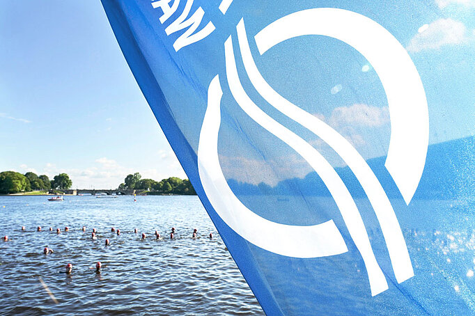 Eine Fahne mit dem Logo von HAMBURG WASSER vor der Alster, in der gerade Triathleten schwimmen