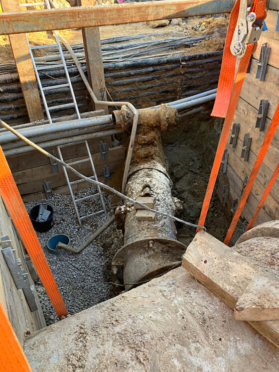Gezeigt wird ein Blick in die Baugrube, die durch Spundwände abgestützt ist. Man sieht ein großes altes Rohr, das unter kleineren Rohren durchläuft.