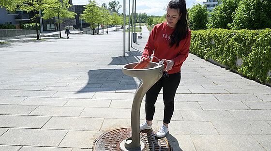 Eine junge Frau füllt ihre Wasserflasche im Sonnenschein an dem neuen Trinkwasserbrunnen auf. Im Hintergrund ist der Inselpark zu sehen.
