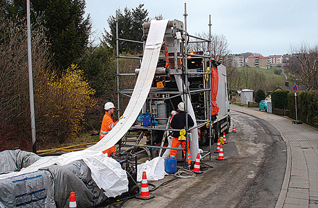 Ein weißer Schlauch liegt auf einem langen Förderband, das auf einem LKW aufliegt. Von hier wird er in den Kanal eingebracht.