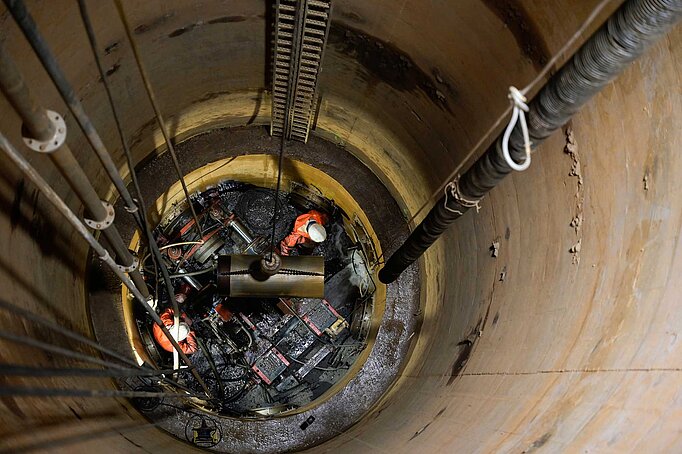 Blick in den Horiontalfilterbrunnen von oben, zu sehen sind Mitarbeiter in einem tiefen Schacht, die mit einem Bohrkopf neue Stränge ziehen. 