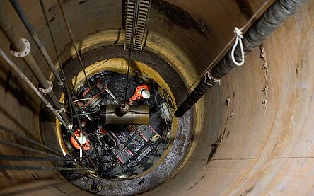 Blick in den Horiontalfilterbrunnen von oben, zu sehen sind Mitarbeiter in einem tiefen Schacht, die mit einem Bohrkopf neue Stränge ziehen. 