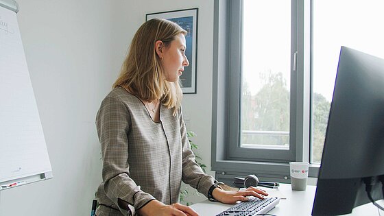 Eine junge Frau steht vor dem Computerbildschirm am Schreibtisch und gibt Daten ein.
